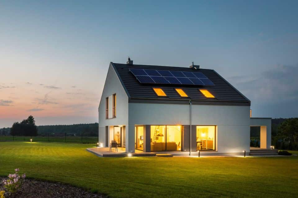 Modernes zweistöckiges Haus mit großen Fenstern, beleuchteten Innenräumen, die über effiziente Stromtarife mit Strom versorgt werden, und Solarmodulen auf dem Dach, umgeben von einem gepflegten Rasenhof in der Abenddämmerung.